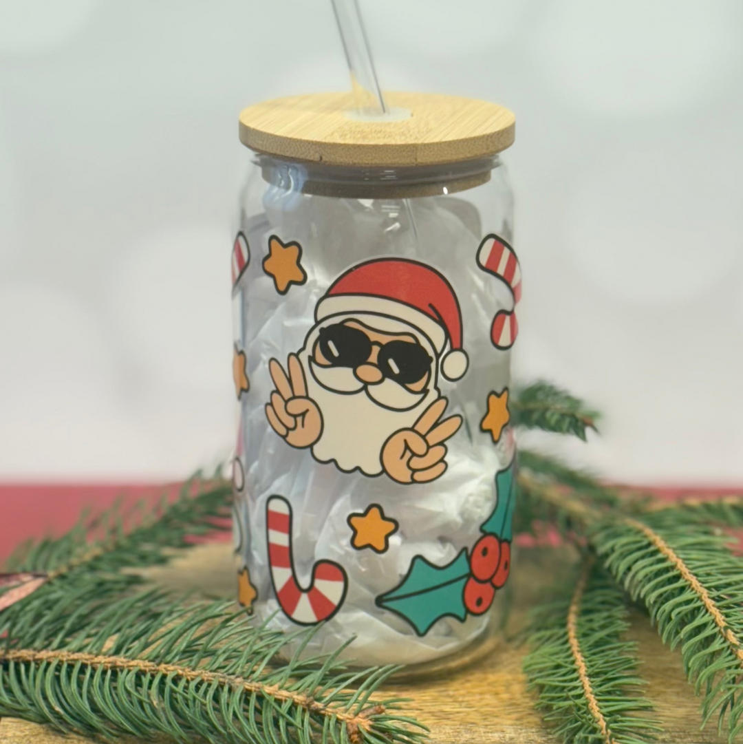 Glass jar with Santa Claus design, wooden lid, and straw on a light background with greenery.