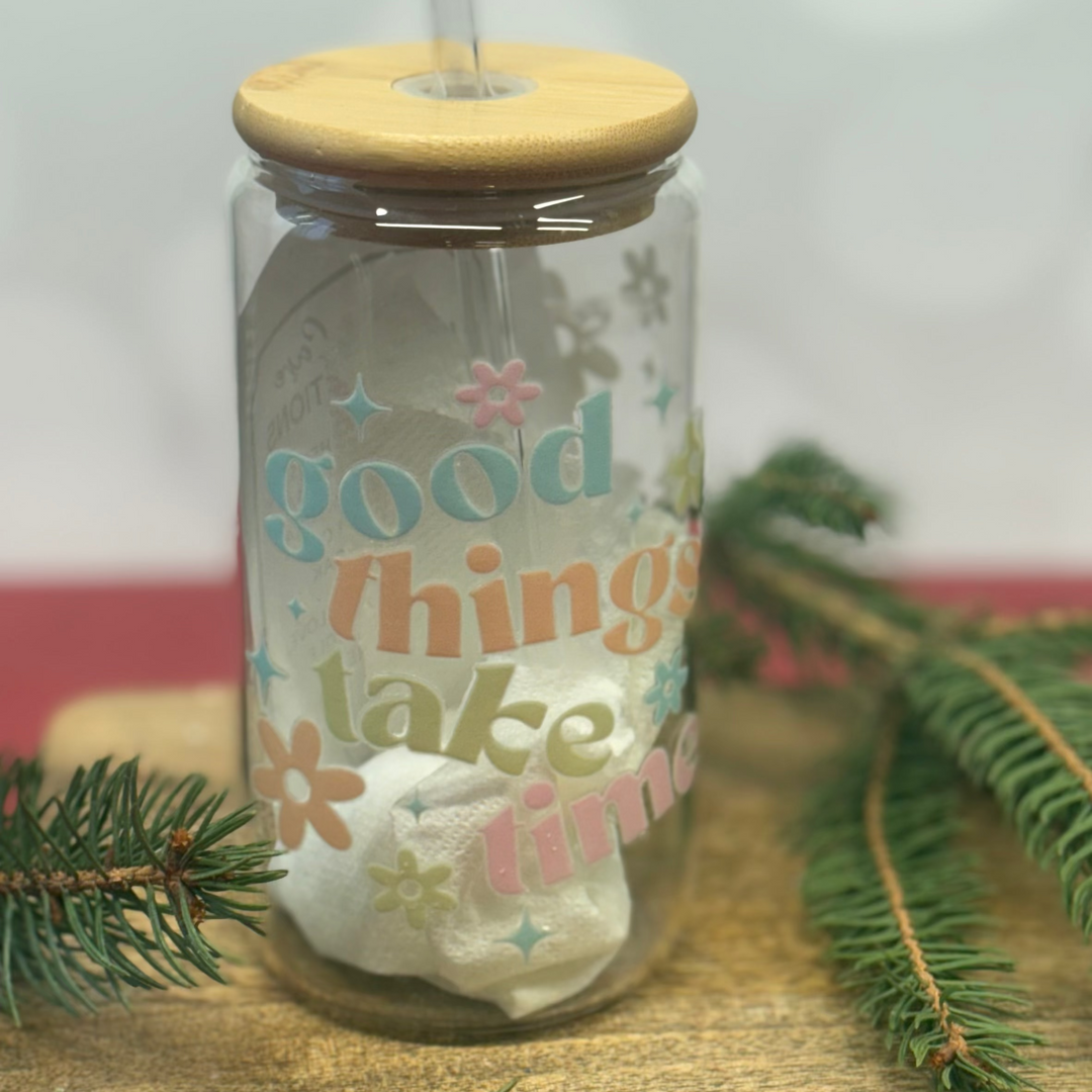Glass tumbler with wooden lid and straw, featuring 'good things take time' text, on a wooden surface with greenery.