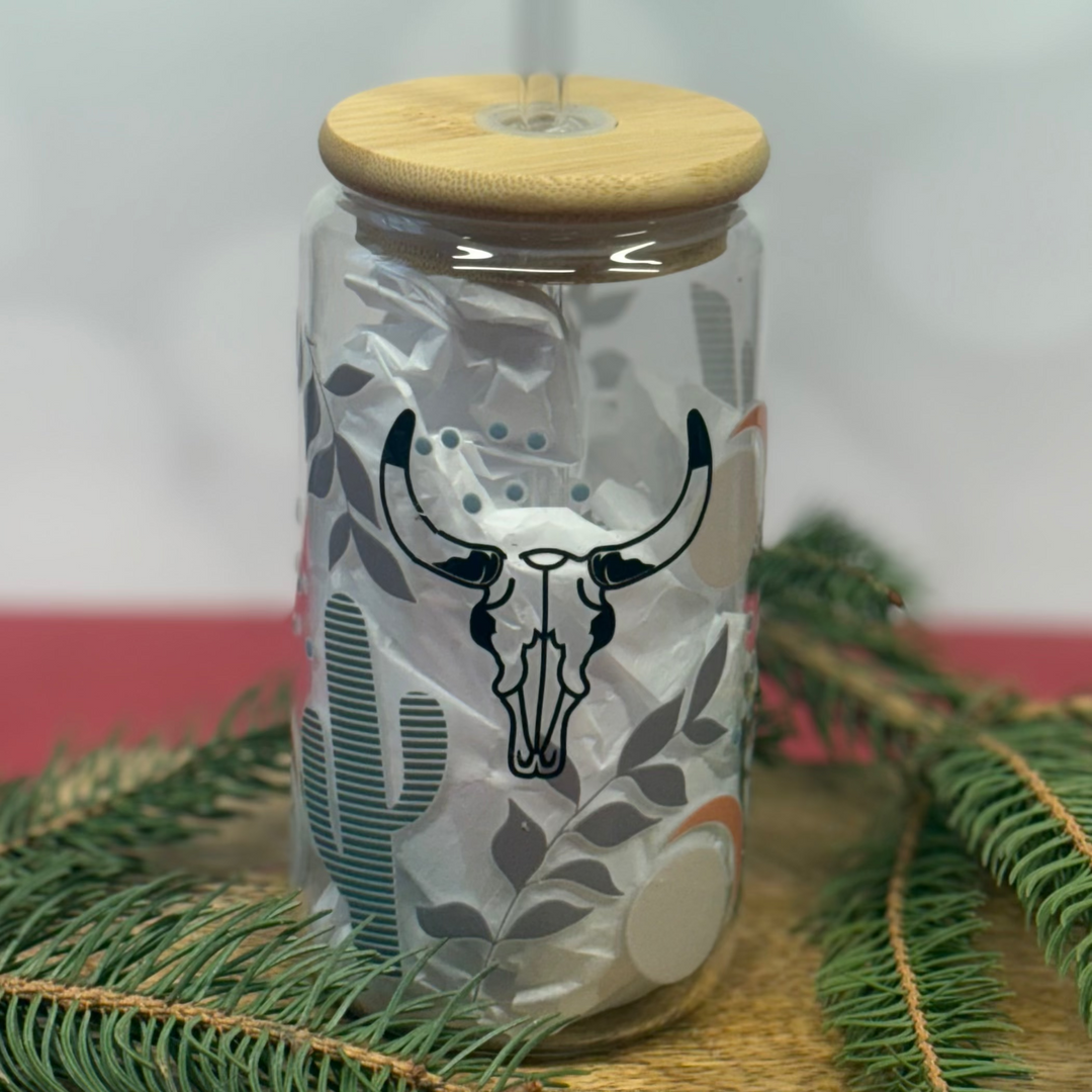 Clear glass tumbler with wooden lid and deer skull design, surrounded by greenery on a white background.