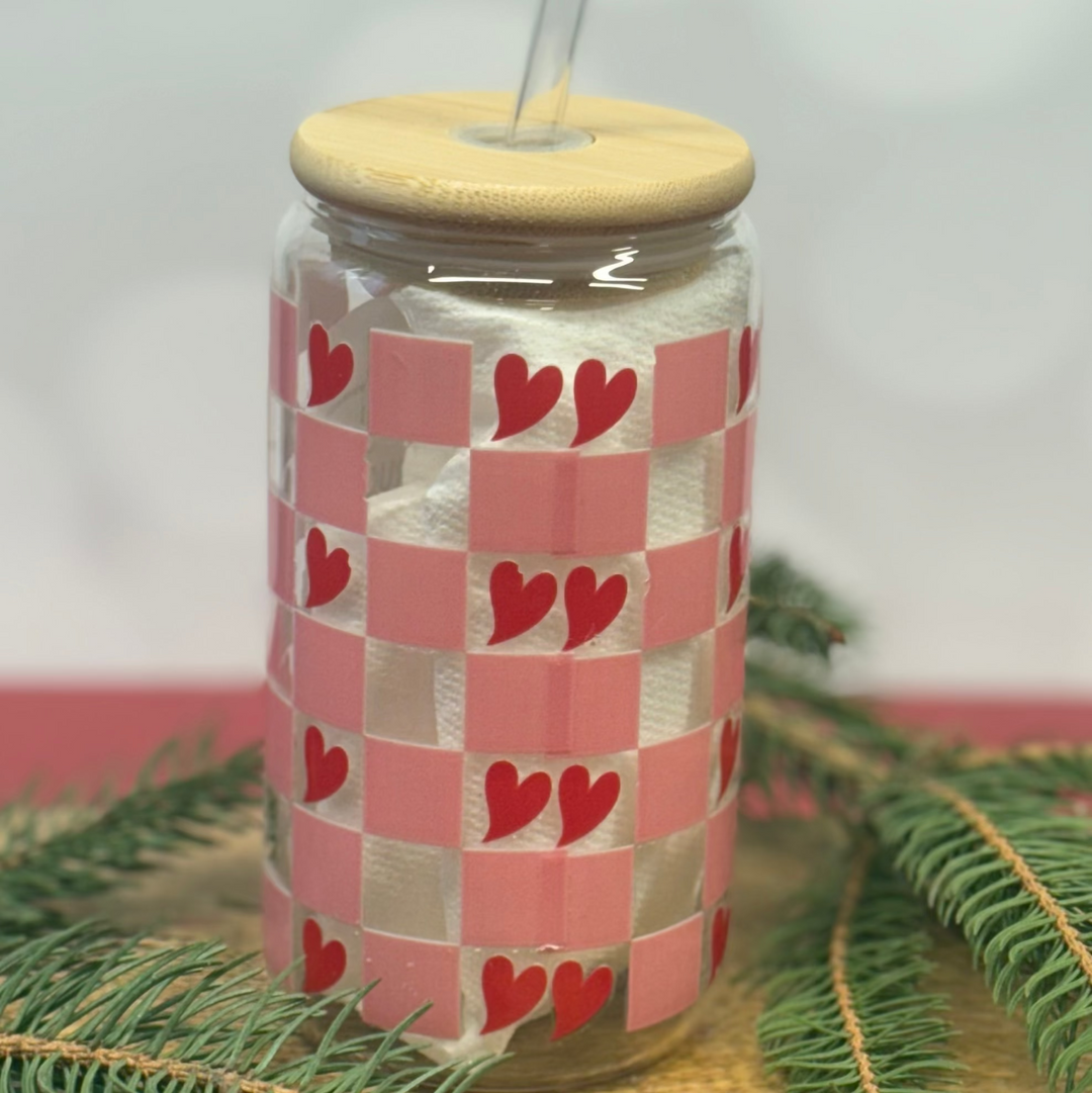 Glass tumbler with a checkered pattern and heart designs, placed on a wooden surface with greenery.